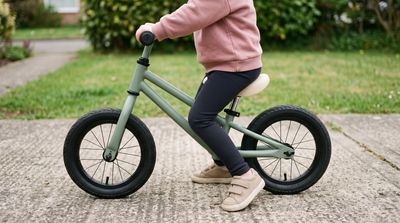 A toddler seated on a plain green balance bike with both feet flat on the pavement, the saddle just below hip height